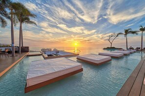 A rooftop infinity pool with sun loungers and a view of the sunset over the ocean.