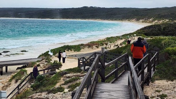 Une promenade en bois qui mène à une plage où l’eau est d’un bleu limpide et le rivage est sablonneux.
