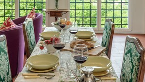 Mesa de comedor dispuesta con platos, vasos y cubiertos, rodeada de sillas con tapicería estampada, y vista a la vegetación a través de ventanas grandes.