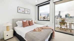 Een moderne hotelkamer met een groot bed, uitzicht over de stad, een balkon met een stoel en een ingelijste foto aan de muur.