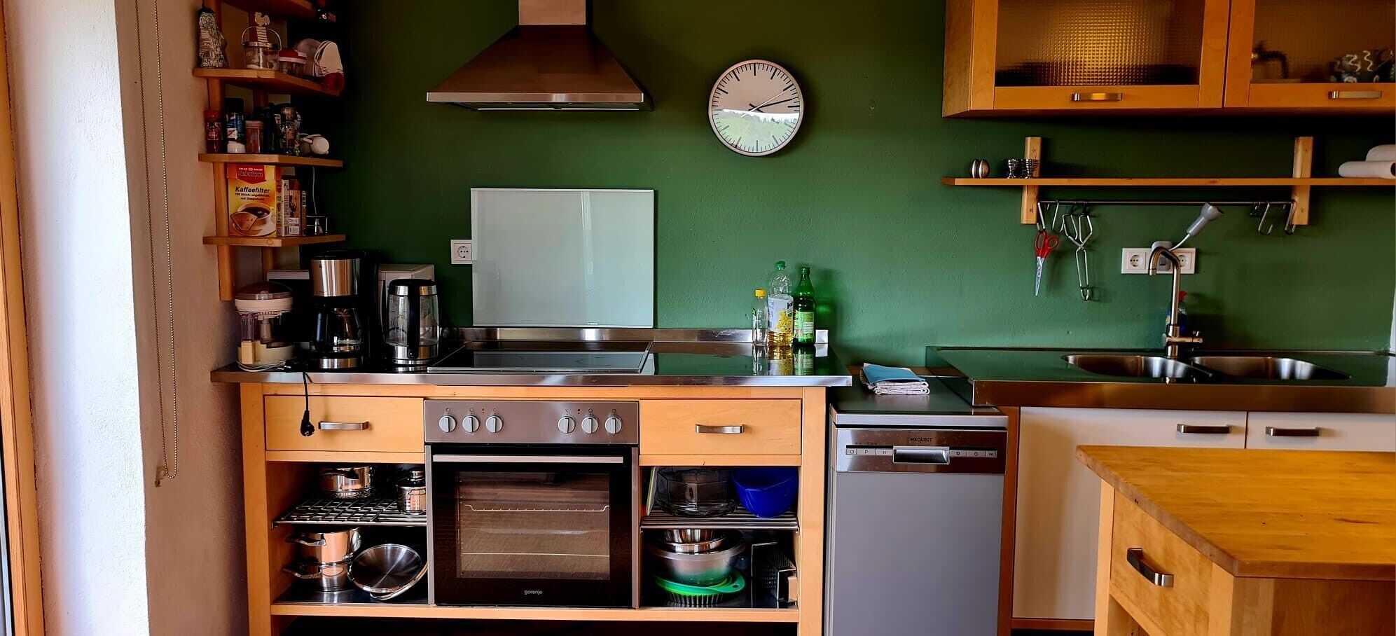 Una cocina compacta con estufa, horno y lavavajillas, armarios de madera y un reloj en la pared.