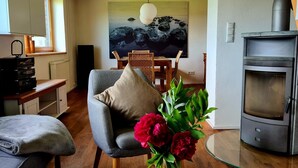 Uma sala de estar com sofá, poltrona, mesa com um vaso de flores, um quadro na parede e uma lareira a lenha.