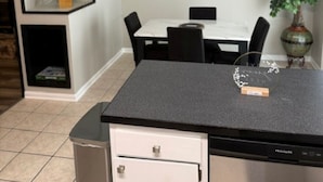 Una cocina compacta con refrigerador, encimera, mesa de comedor con sillas, ventilador de techo, planta en maceta y un cuadro abstracto colgado en la pared.