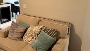 Un sofá color beige con cojines estampados, un cuadro con paisaje de bosque, y un comedor con sillas y una mesa.