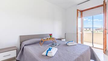 Un dormitorio moderno con cama, mesitas de noche, una bandeja con productos para el desayuno y vistas a la ciudad a través de un ventanal grande.