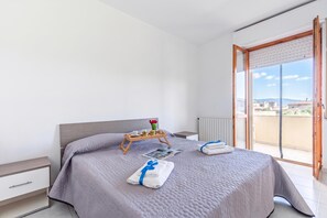 Un dormitorio moderno con cama, mesitas de noche, una bandeja con productos para el desayuno y vistas a la ciudad a través de un ventanal grande.