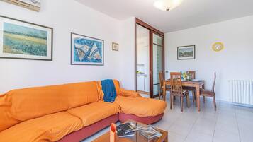 Una sala de estar con un sofá modular, una mesa de comedor con sillas, y una cocina visible a través de una puerta abierta.