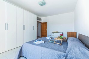 A bedroom with a bed, a bedside table with a tray of items, a wardrobe, and a door leading to another room. - Casa Gialla (Alghero)