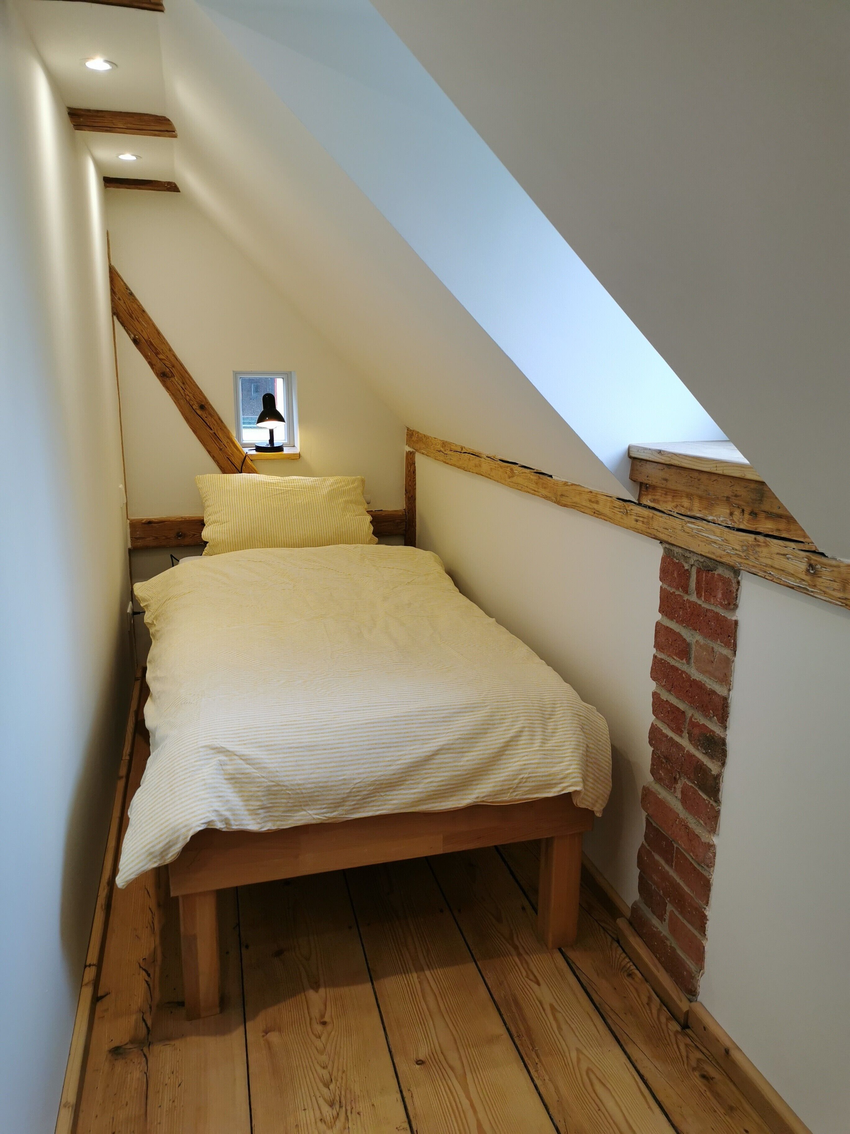 Une petite chambre mansardée, chaleureuse et accueillante, avec un plafond en pente, des poutres en bois, un mur en briques et un lit simple recouvert d’une courtepointe à rayures.