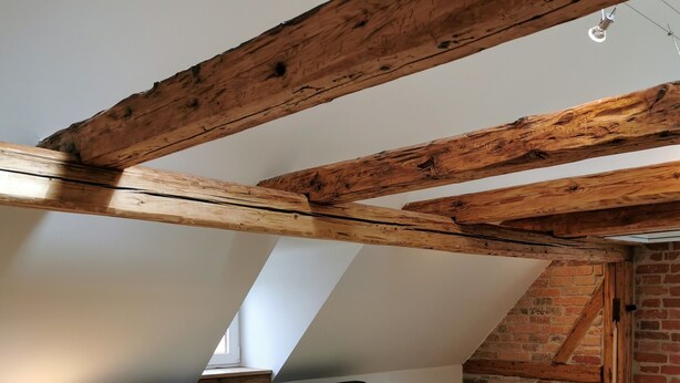 A bedroom with a sloped ceiling, exposed wooden beams, a bed with a dark headboard, a bedside table with a lamp, and a brick wall. - Holiday Home 'Alte Schlosserei' with Private Terrace, Private Garden and Wi-Fi (Presseck)