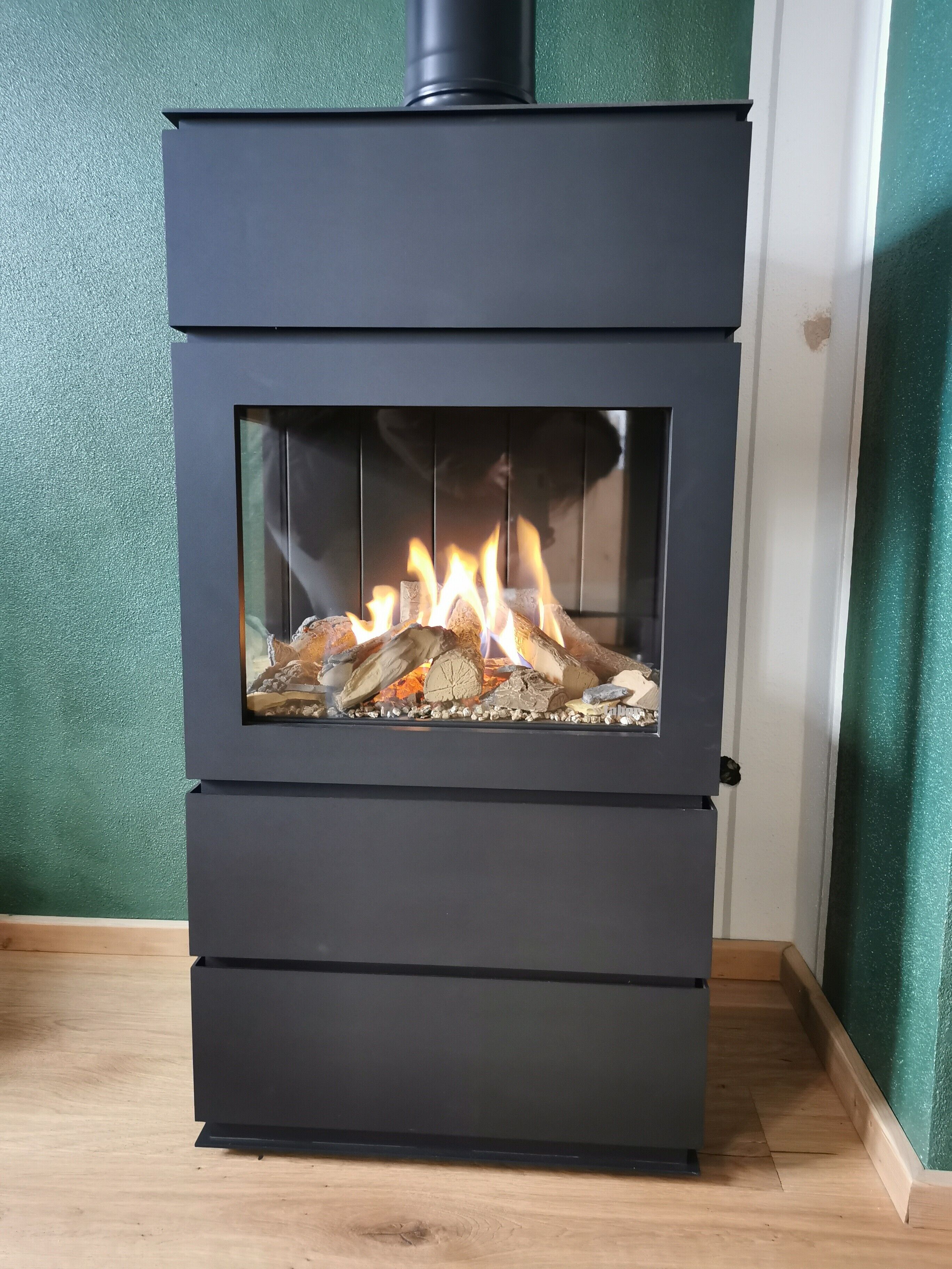 A black wood-burning stove with a glass front, set against a green wall.