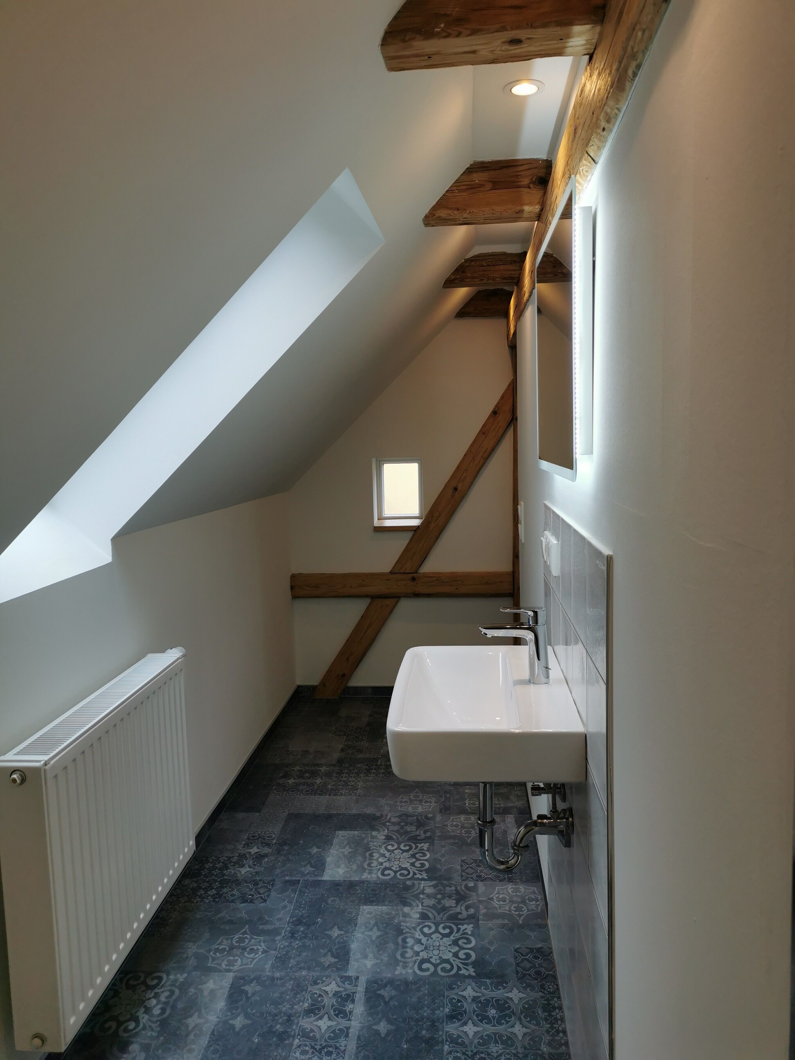 A narrow, sloped hallway with a skylight, a white radiator, a sink with a mirror, and patterned floor tiles.