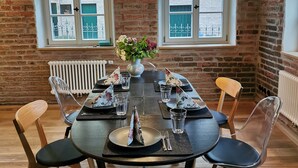 Un comedor con una mesa redonda, seis sillas, una lámpara colgante y paredes de ladrillo con ventanas.