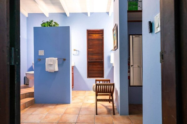 A bathroom with a blue wall, a wooden door, a white towel, a wooden bench, and a window with blinds.