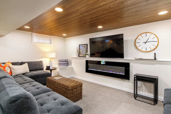 A modern living room with a grey sectional sofa, a flat-screen TV mounted on the wall, a fireplace, and a clock.