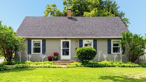 A single-story house with a grey roof, a front door, and a small front yard with a lawn and a few potted plants.