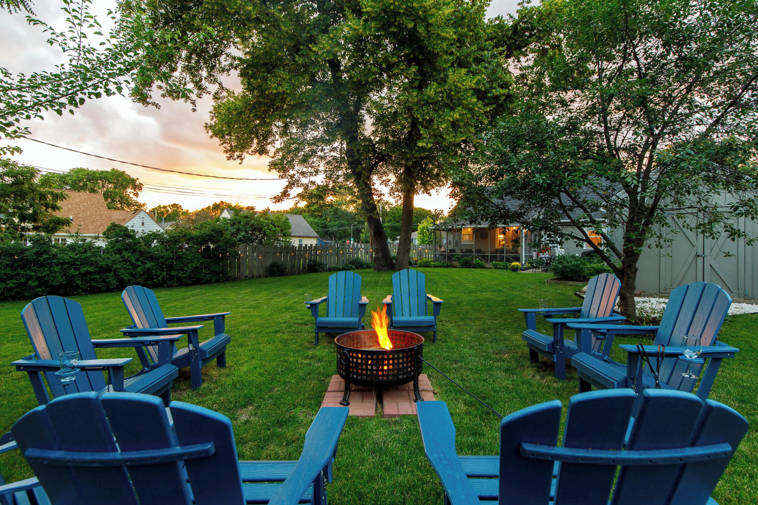 A backyard with a fire pit surrounded by blue Adirondack chairs.