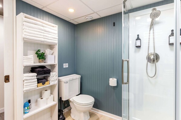 A bathroom with a toilet, a glass-enclosed shower, and shelves with towels and toiletries.