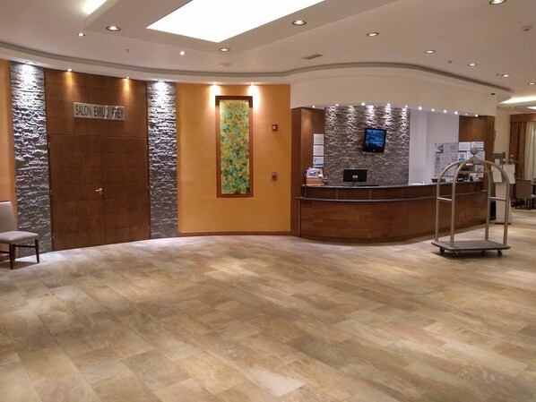 Un área de recepción con una pared de acento de piedra, un mostrador de recepción, una pantalla digital y una zona de estar con una silla.