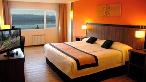 A hotel room with a large bed, two bedside tables with lamps, a TV on a stand, and a window with a view of mountains and a lake.