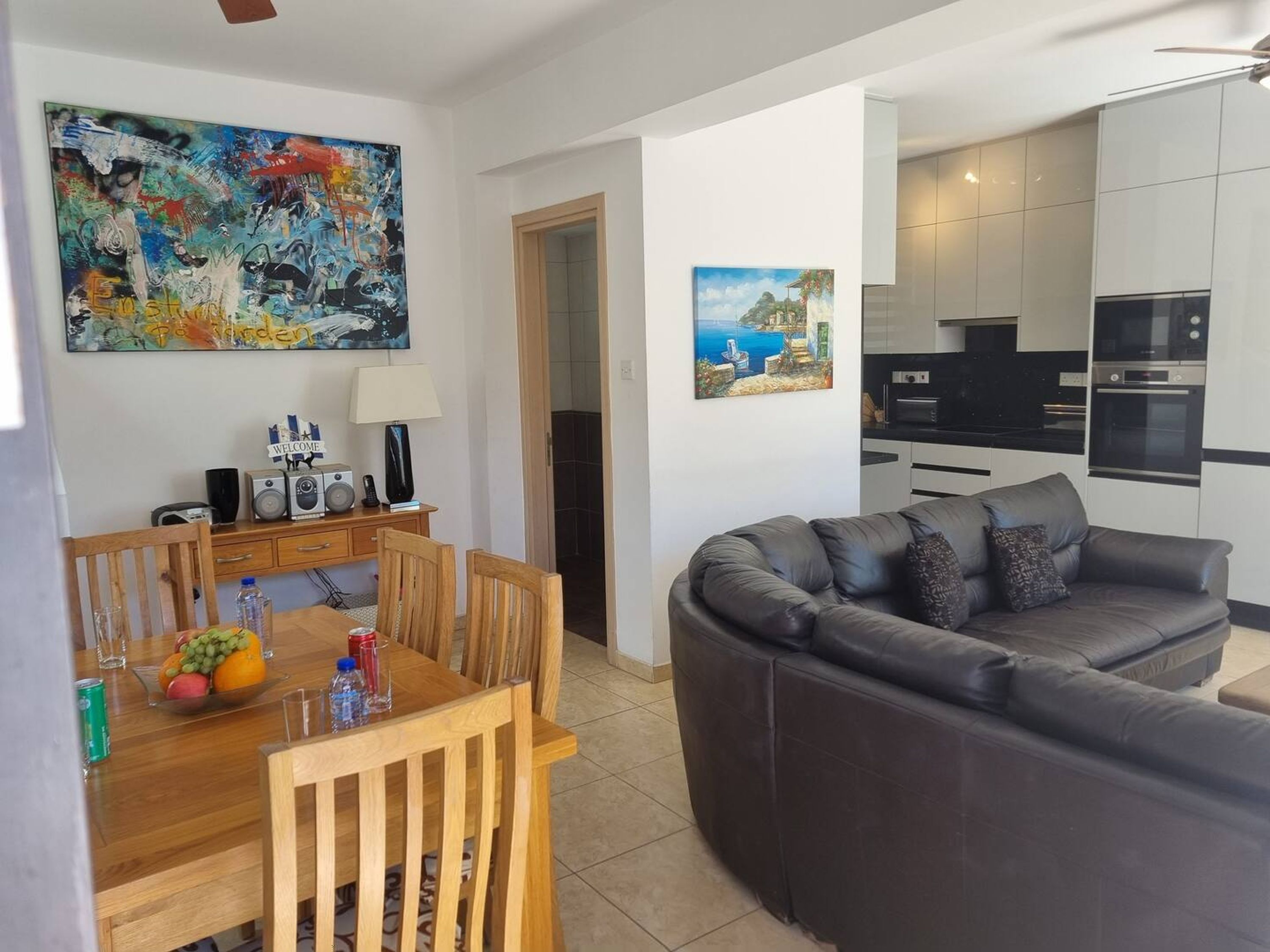 A modern living room with a leather sofa, a wooden dining table with fruit, and a kitchen in the background.