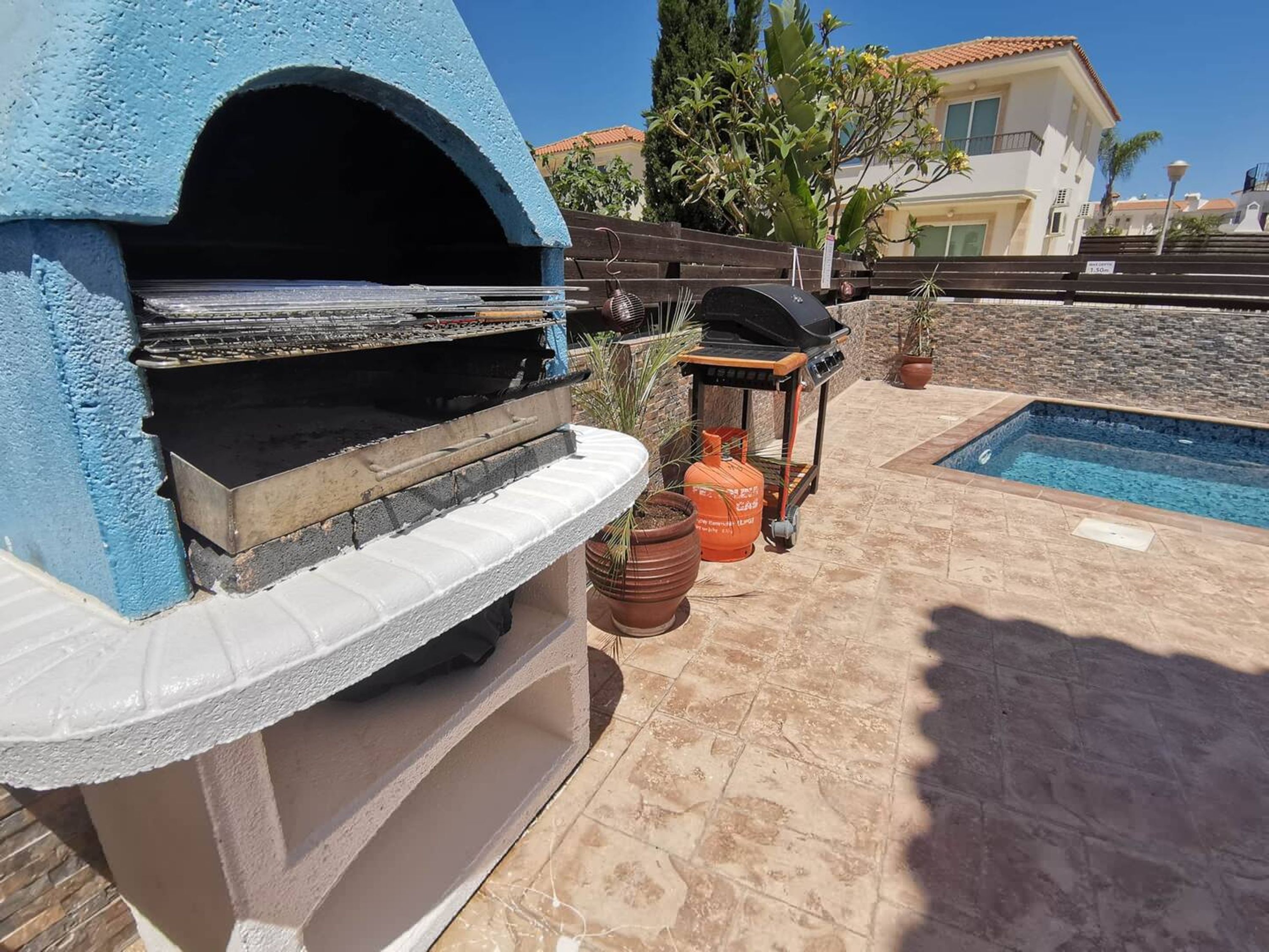 Un horno de pizza azul al aire libre, una parrilla de barbacoa y una zona junto a la piscina con plantas en macetas.