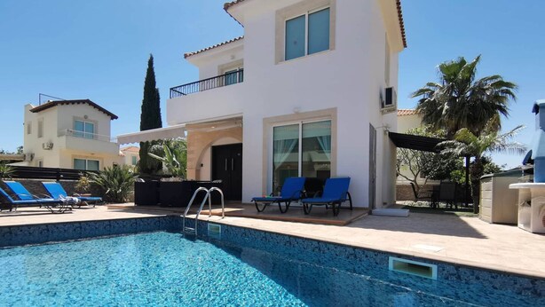 A swimming pool with blue lounge chairs and a white building with a balcony. - Ennazuse - Beautiful 2 Bedroom Villa in Pernera (Paralimni)