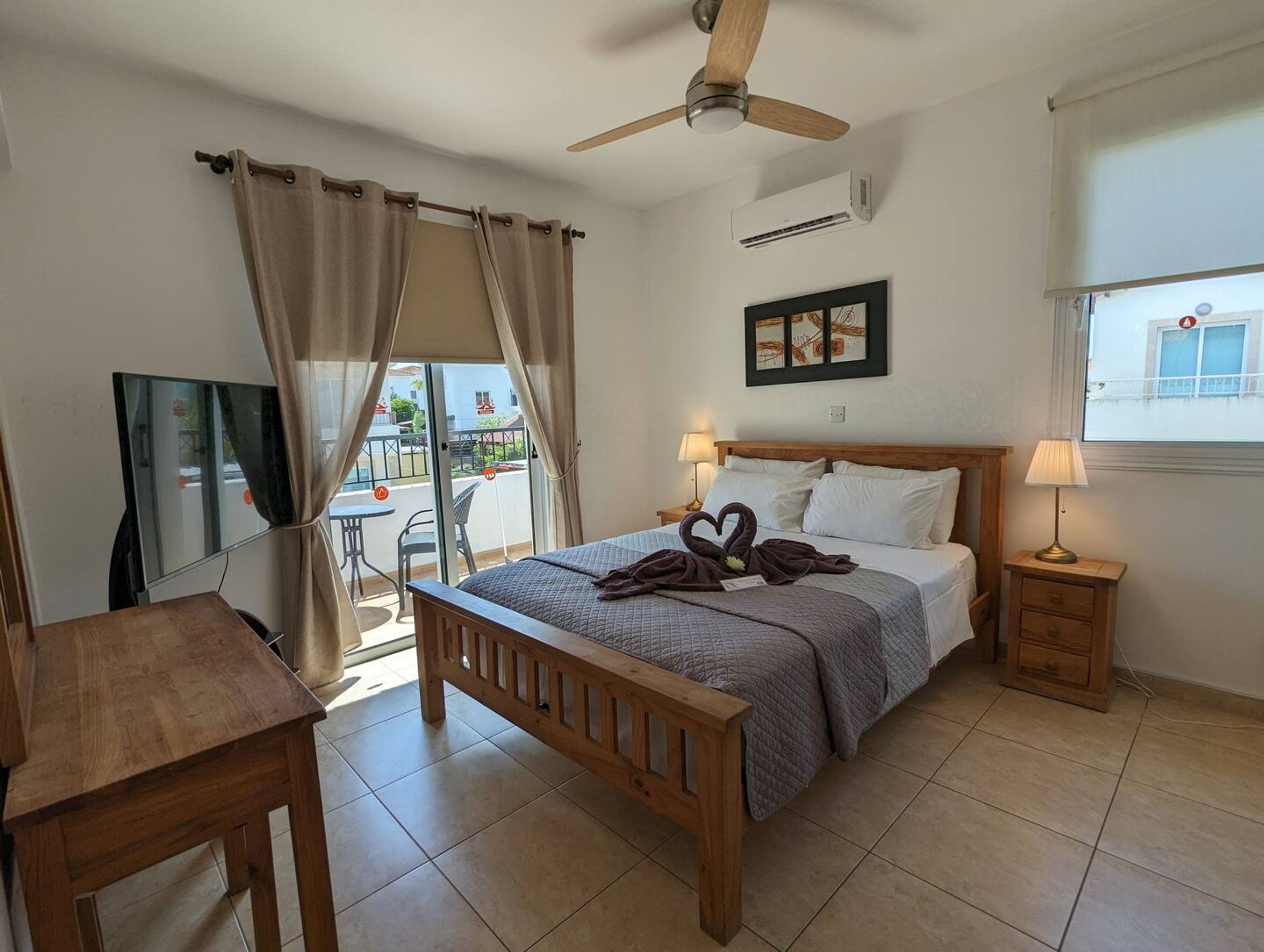 A bedroom with a bed, bedside tables, a ceiling fan, an air conditioning unit, a window with a view, and a desk.