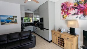 A modern living room with a black leather sofa, a wooden cabinet with wine glasses, a ceiling fan, a kitchen with white cabinets, and a colorful abstract painting. - Ennazuse - Beautiful 2 Bedroom Villa in Pernera (Paralimni)