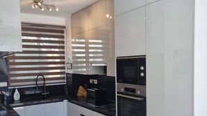 A modern kitchen with white cabinets, a black countertop, and a built-in oven. There is a window with blinds, a ceiling fan, and a black leather sofa in the living area. - Ennazuse - Beautiful 2 Bedroom Villa in Pernera (Paralimni)