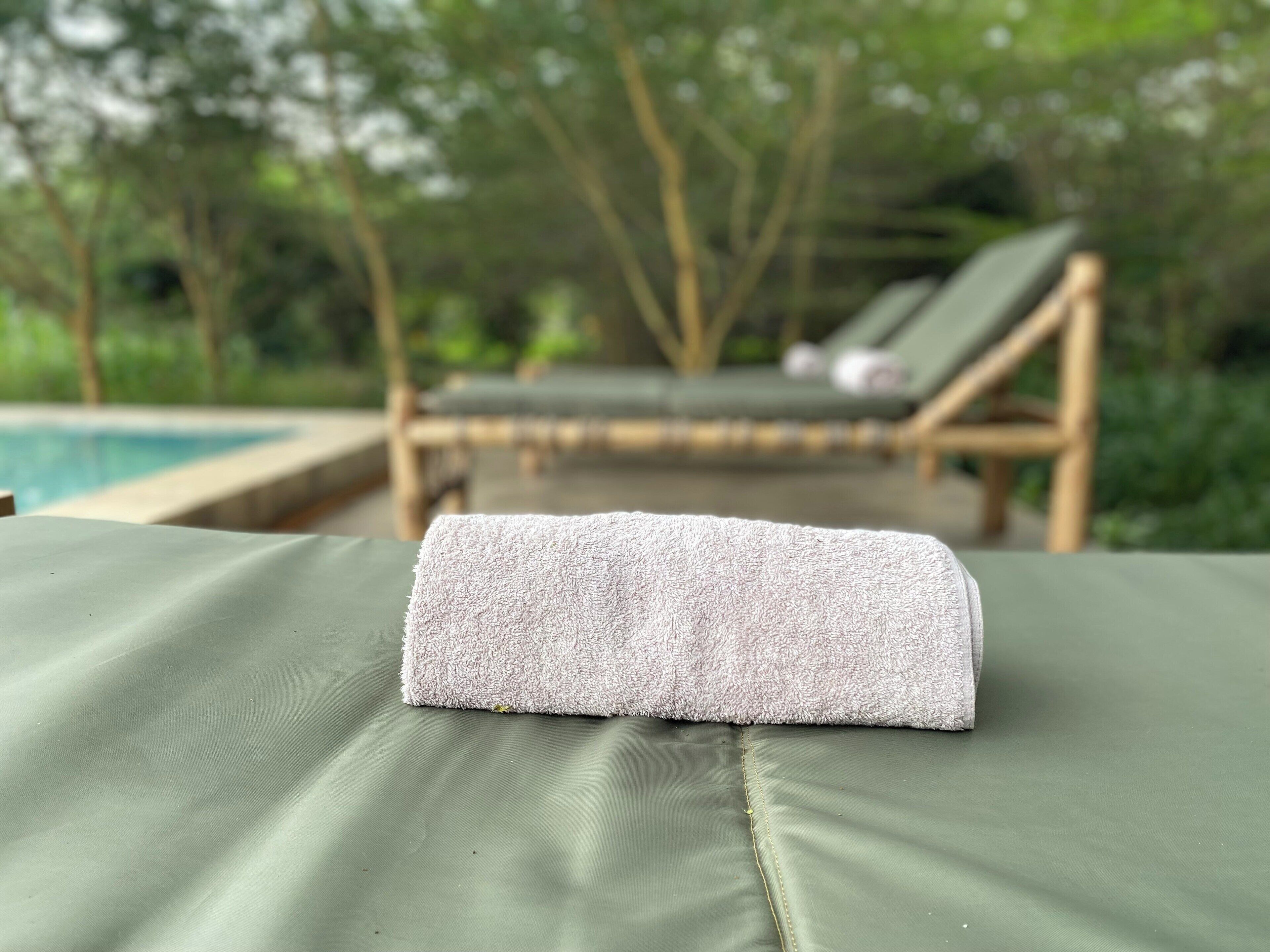 Un fauteuil de jardin vert avec une serviette blanche posée dessus, situé près d’un espace de détente au bord de la piscine, avec un arrière-plan flou composé d’arbres et d’un autre fauteuil de jardin.