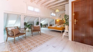 A spacious loft bedroom with a high ceiling, exposed wooden beams, a large bed, a sofa, and a dining area with wicker furniture.