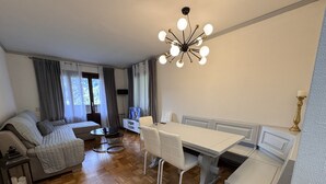 A modern living room with a sofa, a dining table with chairs, a TV, and a chandelier. - 012 Trilocale Pinzolo (Pinzolo)