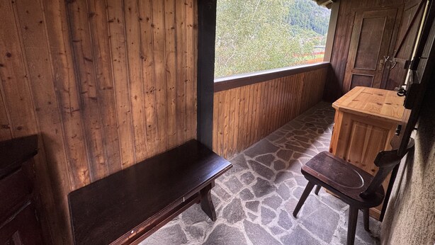A wooden cabin with a stone floor, a bench, a wooden table, and a window offering a view of a forested area. - 012 Trilocale Pinzolo (Pinzolo)