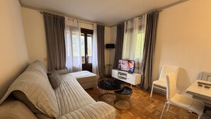 A modern living room with a sectional sofa, a small round table, a TV on a white cabinet, and a white chair. - 012 Trilocale Pinzolo (Pinzolo)