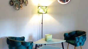 Et værelse med et fiskebensmønstret trægulv, to smaragdgrønne polstrede stole, et lille bord, en gulvlampe og en cirkelformet lampe monteret på væggen.