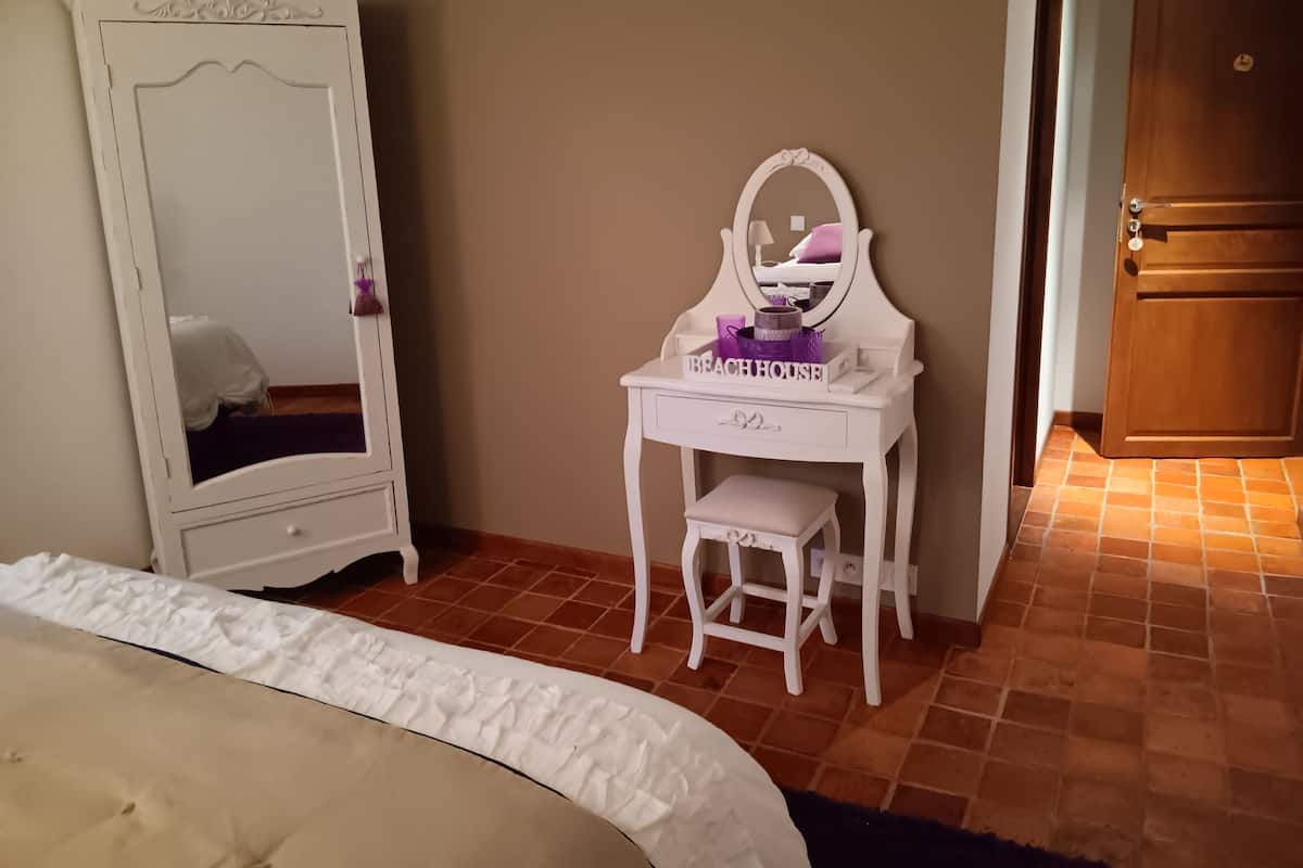 Ein Schlafzimmer mit weißem Schminktisch, einem Spiegel, einem kleinen Hocker und einem Bett mit weißer Daunendecke.