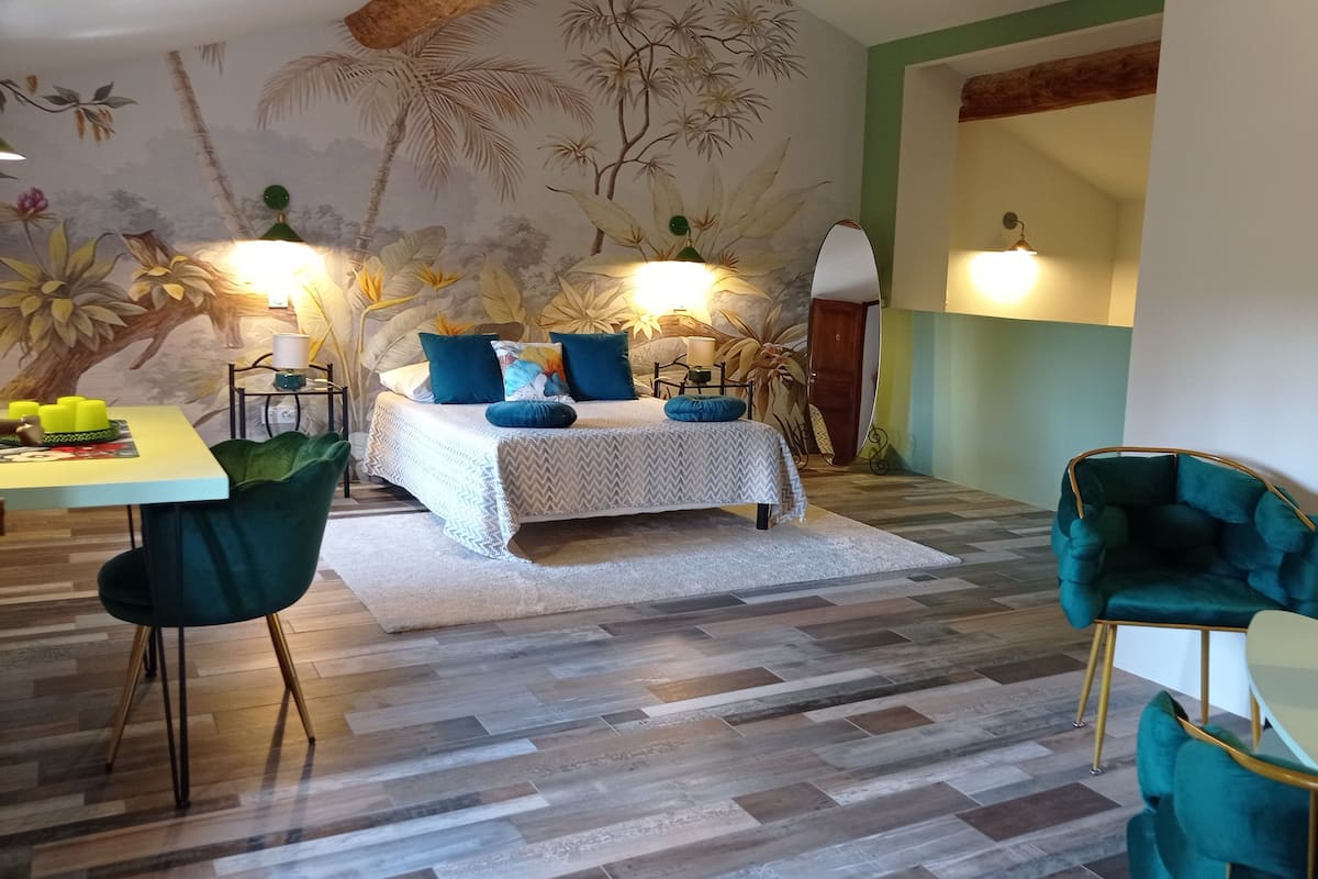 Ein Schlafzimmer mit einem Bett, zwei Stühlen, einem Tisch und einer Wand mit einem tropischen Wandgemälde.