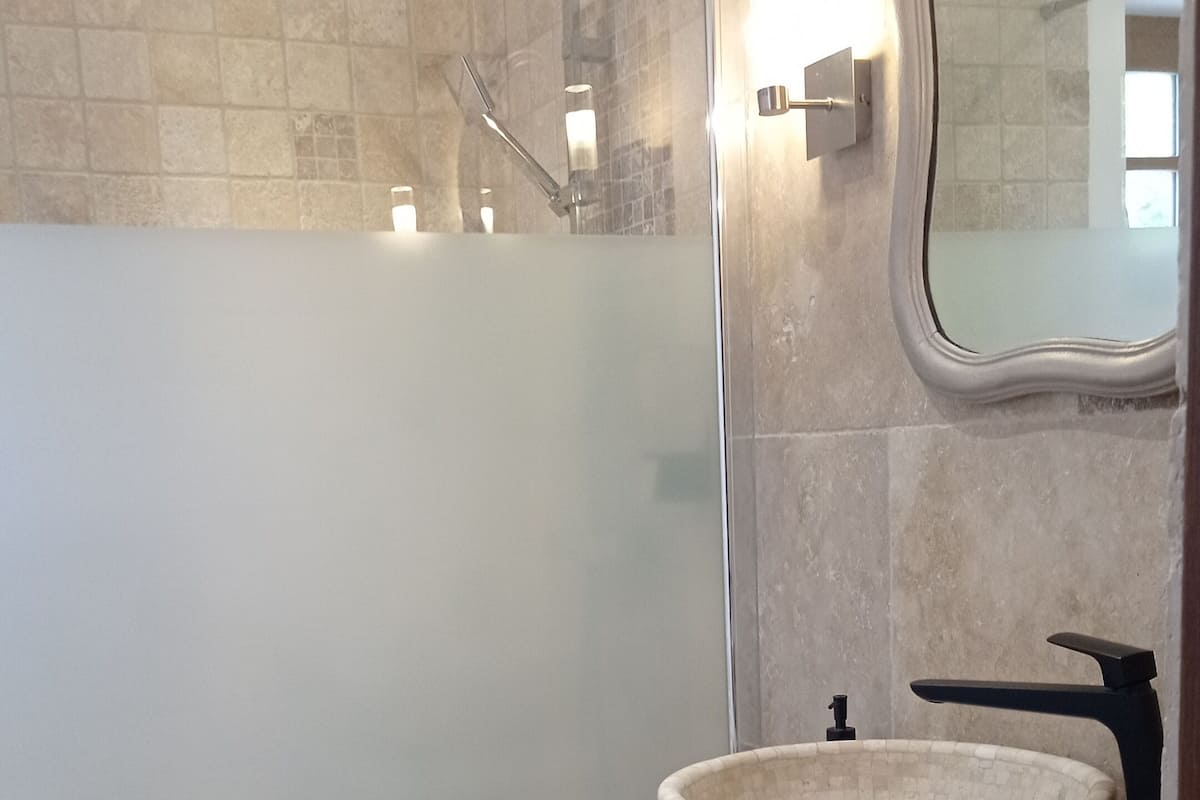 Ein modernes Badezimmer mit einer gläsernen Duschkabine, einem runden Waschbecken und einem blauen Handtuch sowie einem Spiegel mit dekorativem Rahmen.