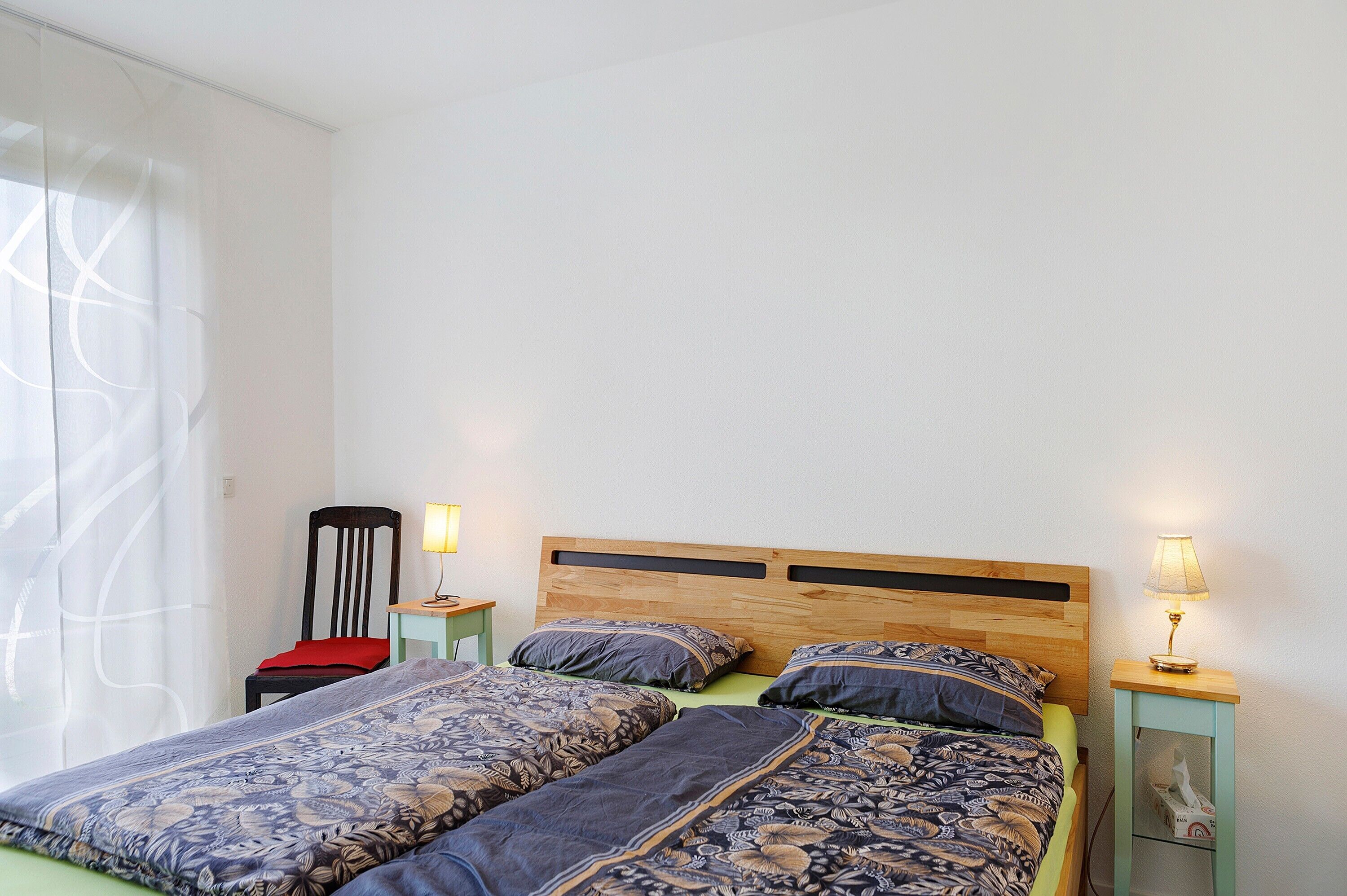 Een slaapkamer met een bed, twee nachtkastjes, een stoel en een raam met gordijnen.