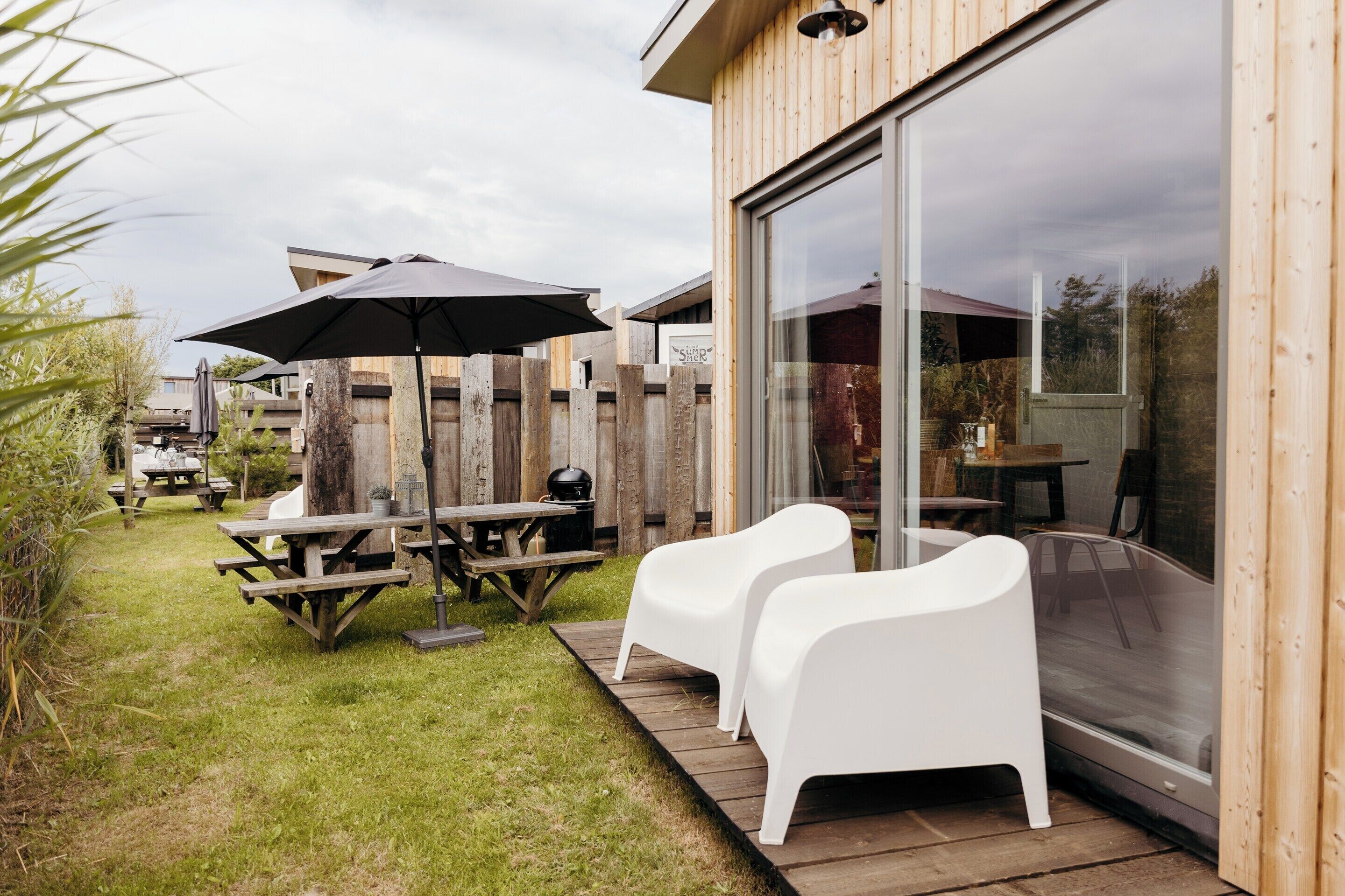 A wooden deck with picnic tables and white chairs, a large umbrella, and a glass door leading to an outdoor area.