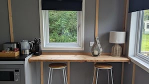 Ett litet kök med ett fönster, två pallar, en kaffebryggare och en lampa.