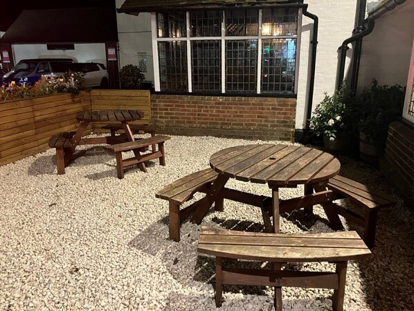 Un conjunto de mesa y sillas de madera sobre grava, con un muro de ladrillo y una ventana al fondo.
