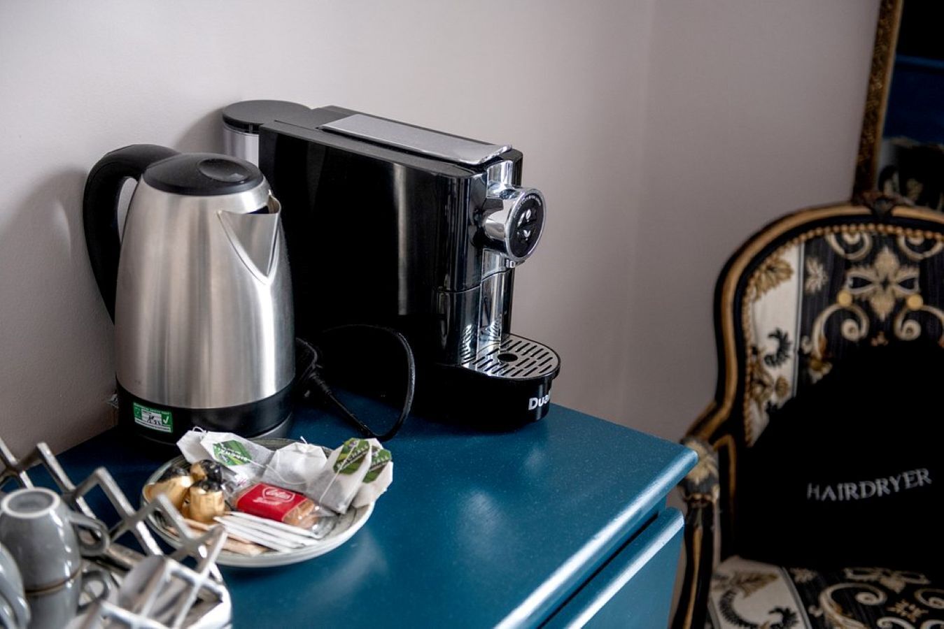 Una cafetera y un secador de pelo sobre una mesa azul, con un plato de comida y cubiertos.