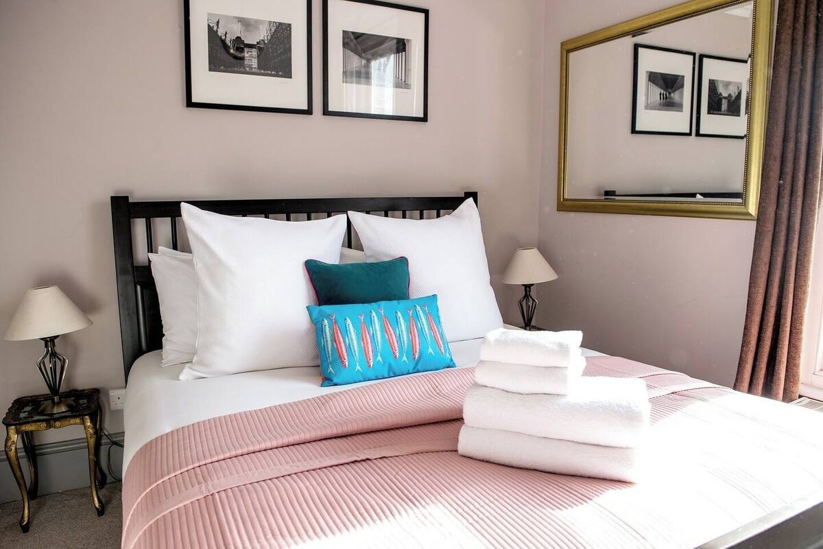 Una cama bien tendida con ropa de cama blanca y rosa, un almohátero color turquesa con estampados de peces, y un espejo con marco dorado que refleja la habitación.
