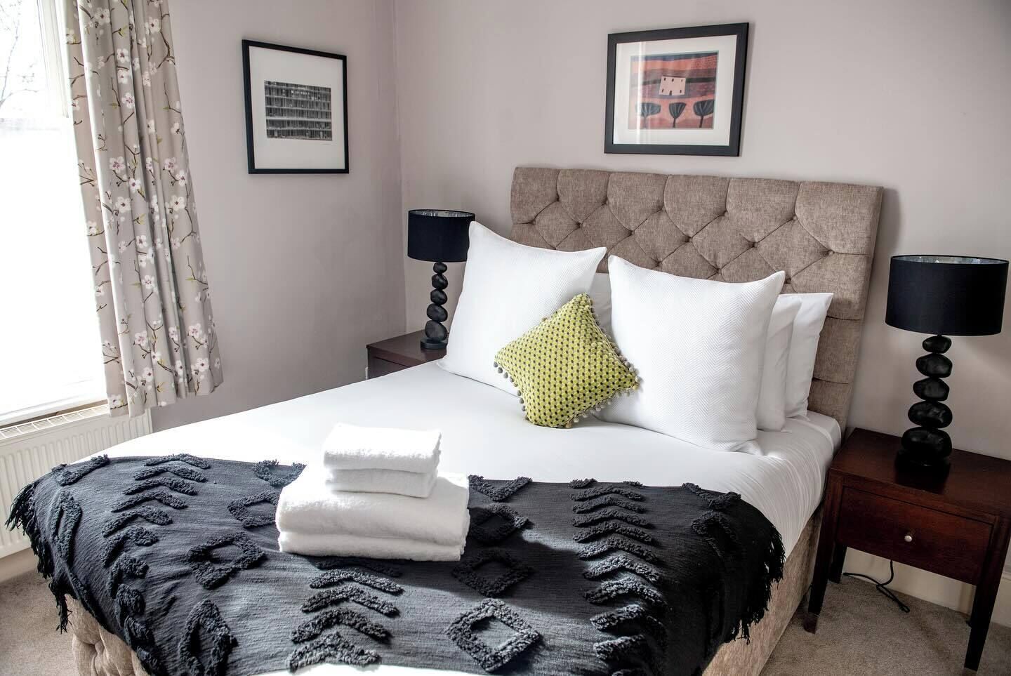 Een net opgemaakt bed met een gestoffeerd hoofdeinde, twee bedlampjes en een raam met bloemen gordijnen.