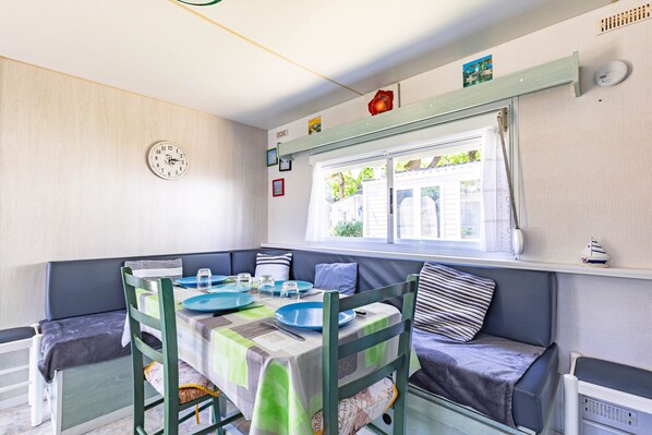 A compact room with a dining table, chairs, a window with curtains, a clock, and a small shelf with decorative items.