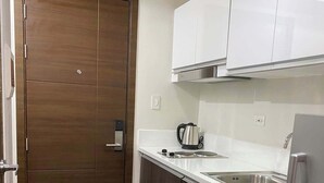 A compact kitchen with a wooden door, white cabinets, a stainless steel sink, and a refrigerator. - Cozy crib @ Air Residences -Makati (Makati)