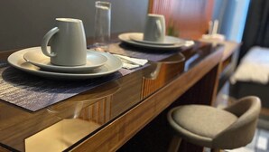A dining area with a wooden bar, two white mugs on saucers, a glass, and a small plate. There are two upholstered chairs and a mirror reflecting the room. - Cozy crib @ Air Residences -Makati (Makati)