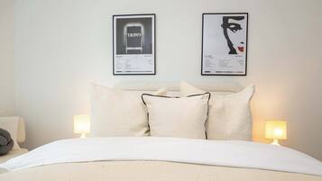 A neatly made bed with white bedding, two wall-mounted lamps, and two framed artworks above the bed.